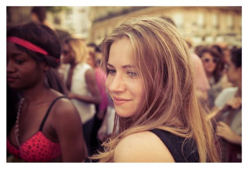 A young woman with long, light brown hair smiles softly as she stands among a crowd of people outdoors, the background slightly blurred to highlight her against the bustling scene. Wearing a black top, she seems to radiate an aura of love while enjoying the lively atmosphere.