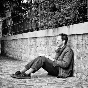 A man sits in quiet méditation on a cobblestone path, leaning against a stone wall and reading a book. Trees and a metal fence line the top of the wall in this serene black-and-white square scene.