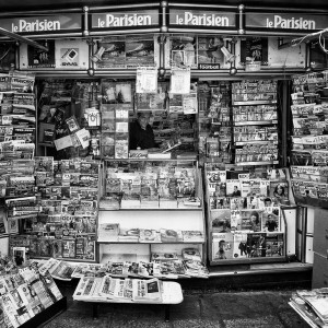 A black-and-white photo of a newsstand filled with newspapers and magazines. A person sits inside, partially obscured by publications. Signs for "Le Parisien" are displayed at the top of the stand.