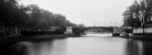 A black and white photo of a serene river, Au fil de l'eau, with a stone-arched bridge spanning across it. Trees line the riverbanks and a few buildings are visible in the background. The calm water reflects the bridge and surrounding scenery.