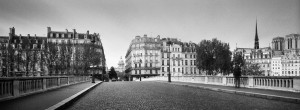 A black and white photo of a cobblestone bridge spanning a river, connecting two sections of a city with classic European architecture. Au fil de l'eau, landmarks and church spires rise in the background, while leafless trees line the sides of the image.