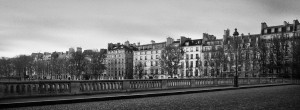 A black-and-white photograph of a European cityscape, showcasing a row of classic, multi-story buildings with numerous windows. Au fil de l'eau, a stone bridge with a balustrade crosses in the foreground, flanked by bare trees, likely taken in the fall or winter season.
