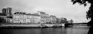 A black and white image of a riverside cityscape, captured "au fil de l'eau," with several multi-story buildings lined along the water. A bridge crosses the river in the background, and a large cathedral with twin towers is visible on the left. Overhanging tree branches frame the right side.