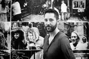 A man with a beard stands in front of a collage of black-and-white street photographs featuring various people and urban scenes. He looks directly at the camera with a neutral expression, embodying the intrigue of each Beautiful One captured behind him.