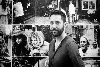 The Beautiful One A man with a beard stands in front of a collage of black-and-white street photographs featuring various people and urban scenes. He looks directly at the camera with a neutral expression, embodying the intrigue of each Beautiful One captured behind him.