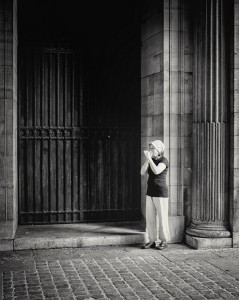 A woman stands alone near a large column and an iron gate, appearing to pray or contemplate, sous les arcades of a dimly lit stone building with cobblestone pavement. The black and white scene captures her quiet solitude.