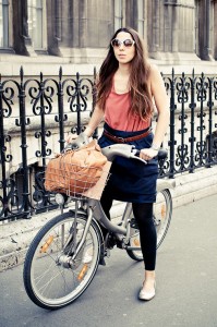 La passagère, wearing sunglasses, a coral top, black skirt, and black leggings, walks her bicycle with a basket holding a tan bag along a city street beside an ornate iron fence.