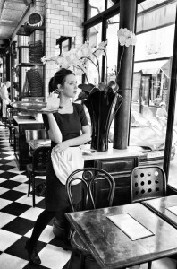 A jolie fleur waitress in a black dress and white apron stands in the café, thoughtfully gazing out the window. The checkered floors, empty tables, and a large vase with orchids add charm to the tranquil scene.