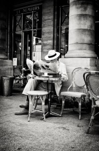 A person wearing a white suit and hat sits alone at an outdoor café table, reading pages of a newspaper. Classic wicker chairs and tall stone columns create a Parisian street scene rich in timeless cultures, all captured in black and white.