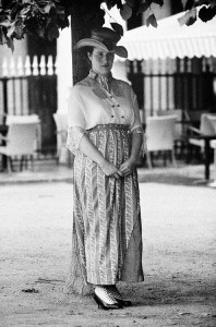 A woman stands under a tree, wearing a wide-brimmed hat and a dress with vertical stripes and lace details. She has a neutral expression and clasps her hands in front of her, embodying the elegance of dames. In the background, there are chairs and tables, suggesting an outdoor seating area where she finds quiet bonheur.