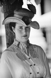 Black and white photograph of a woman from the early 20th century in a patterned dress and a large, ornate hat. She stands in front of a tree, looking calmly at the camera with a neutral expression—a true portrait of bonheur among dames of her era.