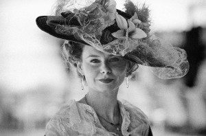 A sepia-toned image of a woman in vintage attire captures the essence of "Les Dames du Bonheur." She wears an elaborate hat adorned with lace, flowers, and netting, complemented by a delicate blouse with lace details. Her long earrings dangle gently as she smiles slightly, against a softly blurred background.