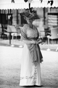 A woman wearing a vintage-style dress and an elaborate hat stands outdoors. The black and white image captures this dame holding a shawl, with chairs and a building's porch with columns in the background. The image exudes nostalgic bonheur, evoking a historical feel.