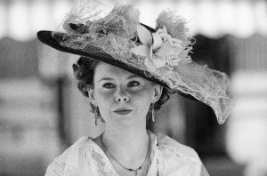 Black and white image of a person wearing a vintage, wide-brimmed hat adorned with flowers and lace. They have curly hair, earrings, and a delicate necklace. The blurred background draws focus to the person's serene expression, exuding an air of timeless bonheur akin to the elegance of dames from another era.