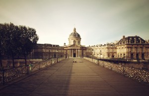 A wide wooden bridge, the Pont des Arts, leads to the grand domed Institut de France with classical architecture, flanked by historic structures. Trees and railings covered with padlocks line the bridge under a clear sky.