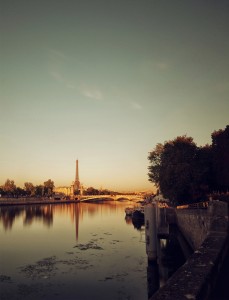 A peaceful view of the Eiffel Tower at sunset, reflected in the calm waters of the Seine River, with trees and walkways lining the riverbank under a clear sky—capturing the charm often found au petit matin.