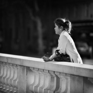 A woman leans on a stone balustrade, gazing into the distance with an air of mélancolie. Dressed in a light-colored top, she holds a bag and wears her hair in a ponytail. The scene is in black and white, with a blurred background, creating a contemplative and serene atmosphere.
