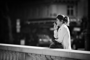 A black-and-white photo showing a woman standing outdoors, leaning against a stone railing. She is dressed in a light-colored coat, has a ponytail, and holds a cigarette in her right hand, looking contemplatively into the distance with an air of mélancolie. The urban background is blurred.