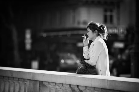 A black-and-white photo showing a woman standing outdoors, leaning against a stone railing. She is dressed in a light-colored coat, has a ponytail, and holds a cigarette in her right hand, looking contemplatively into the distance with an air of mélancolie. The urban background is blurred.