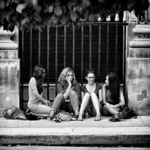 Four young women sit on a sidewalk in front of a metal fence, chatting and relaxing together despite the chaleur. Bags rest beside them as they enjoy each other’s company. The black-and-white photo captures a carefree moment.