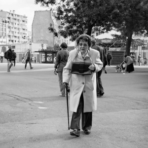 An elderly woman wearing a coat, sunglasses, and a delicate pendentif walks with a cane in an urban square. Several people are in the background, along with trees, buildings, and a carousel. The photo is in black and white.