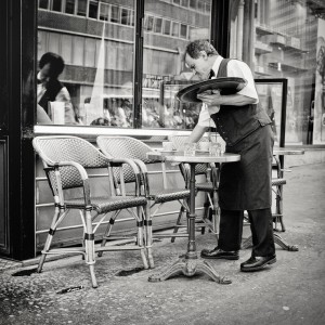 A waiter in a dark apron, with humilité, clears cups from an outdoor café table on a city sidewalk. Two empty woven chairs are beside the table, with reflections of people in the café window behind.
