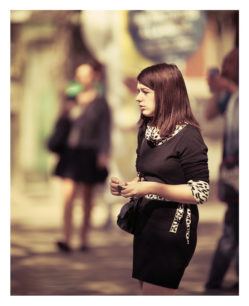 A woman with shoulder-length dark hair is standing outdoors at the 2012 Techno Parade, looking to her left. She is wearing a black dress with a leopard-print belt and scarf, and appears to be holding something in her hands. The background is slightly blurred, with out-of-focus figures and buildings.