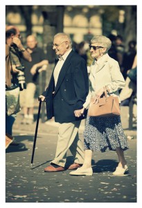 An elderly couple walks hand in hand on a busy street during the 2012 Techno Parade. The man uses a walking stick and wears a dark jacket and light trousers. The woman, in a white jacket, patterned skirt, and sunglasses, carries a handbag. People and confetti are visible in the background.