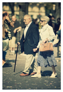 An elderly couple walks hand in hand on a busy street during the 2012 Techno Parade. The man uses a walking stick and wears a dark jacket and light trousers. The woman, in a white jacket, patterned skirt, and sunglasses, carries a handbag. People and confetti are visible in the background.