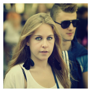 A young woman with long blonde hair, wearing large black earrings and a white top, looks directly at the camera. Behind her, a young man with short hair, sporting black sunglasses and a blue and white shirt from the 2012 Techno Parade, is slightly out of focus.