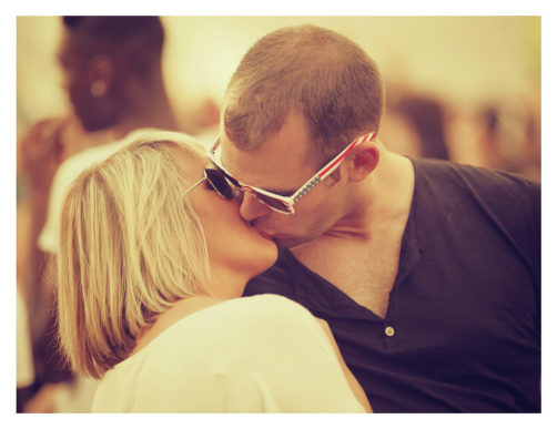 A couple kisses affectionately, both wearing sunglasses. The woman has blonde hair, and the man is wearing a black shirt. The background, reminiscent of a 2012 Techno Parade, is blurred with other people, suggesting an outdoor gathering or event.