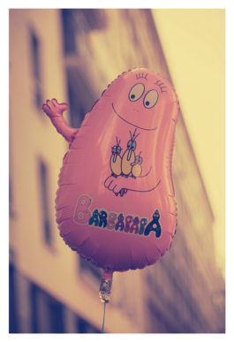 A pink balloon in the shape of a cheerful cartoon character with a round body, large eyes, and a friendly smile. The character has three yellow flowers on its chest and the text "Barbamama" beneath it. The background features an out-of-focus urban scene reminiscent of the 2012 Techno Parade.