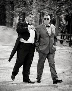 Two men in formal wear, one in a queue de pie tuxedo with tails and the other in a suit and bow tie, walk confidently side by side down a tree-lined path in an outdoor setting. The image is in black and white.