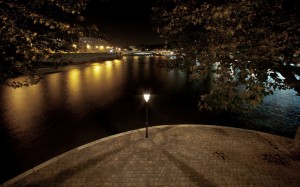 A nighttime view of a cobblestone pathway by the river, lit by a solitary streetlamp. Water reflects the city lights, with trees framing the upper and right sides of the image. The scene is quiet and serene, reminiscent of nuit on tranquil îles, with buildings and a bridge visible in the background.
