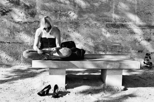 Je ne suis pas de celles qui meurent de chagrin A woman sits cross-legged on a stone bench outdoors, lost in reading or writing on papers. Barefoot, with her shoes set aside and a bag nearby, she seems deep in thought, touched by chagrin as sunlight casts shadows on the wall behind her.