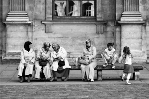 Four women wearing headscarves sit on a bench outside a stone building, engaged in échanges féminins. Nearby, a boy and girl, both casually dressed, sit and stand. The scene appears candid and relaxed.