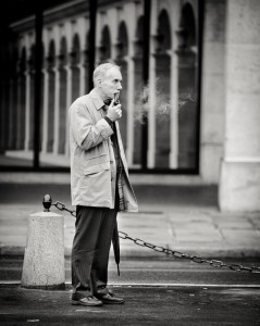 A fière allure, the homme à la pipe stands in a long coat on a city street, looking thoughtful. Black and white tones frame arched windows and a sidewalk lined with stone posts linked by a chain.