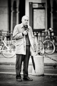 A man with an undeniable allure stands on a city street, homme à la pipe in hand and umbrella by his side. Bicycles are parked behind him, and a blurred map or sign appears in the black-and-white background.