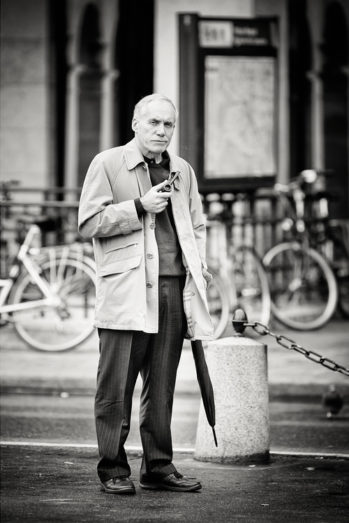 A man with an undeniable allure stands on a city street, homme à la pipe in hand and umbrella by his side. Bicycles are parked behind him, and a blurred map or sign appears in the black-and-white background.