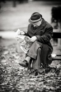 An elderly man in a coat and hat sits on a park bench covered with fallen leaves, rêvant as he reads a newspaper. The black and white photo captures a calm, contemplative moment outdoors.