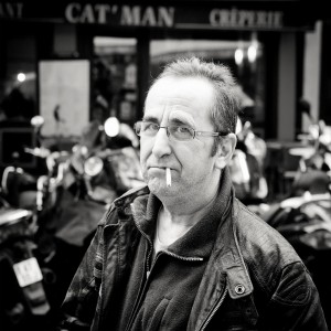 A middle-aged homme with glasses and a cigarette in his mouth stands outdoors in front of the "CAT'MAN" café. Motorbikes are parked behind him, capturing a classic biker vibe in this black and white scene.
