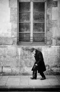 An elderly man in a long coat and cap walks slowly along a sidewalk beside a weathered stone wall with graffiti, the black and white scene capturing le poids des années etched into every step.