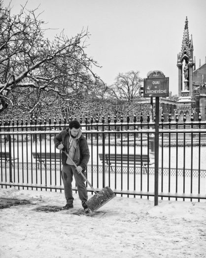 A person shovels neige on a sidewalk beside a wrought iron fence on Quai de l'Archevêché in a snowy landscape. Snow-covered trees and a Gothic architectural structure are visible in the background under the cloudy samedi sky.