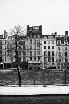 Black and white photo of a row of tall, narrow buildings on a snowy day in Paris. The buildings stand behind a stone wall with bare trees in front, next to a body of water. The scene appears to be cold and quiet, with everything covered sous une couche de neige.