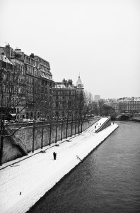 Black-and-white photo of a snowy urban scene by a river. Leafless trees line the walkway parallel to the water, bordered by tall buildings. People walk on the snow-covered path beside the river, with tracks marking the neige. An overcast sky enhances the wintry atmosphere sous Paris' charm.