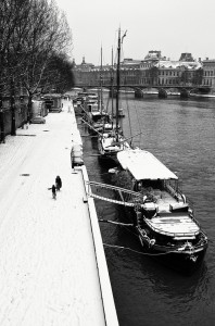 Black and white image of a snow-covered riverside with boats docked along the Seine River in Paris. A person with a dog walks along the snowy path, and trees line the bank to the left. In the background, buildings and a bridge crossing the river are visible, capturing Paris sous la neige.