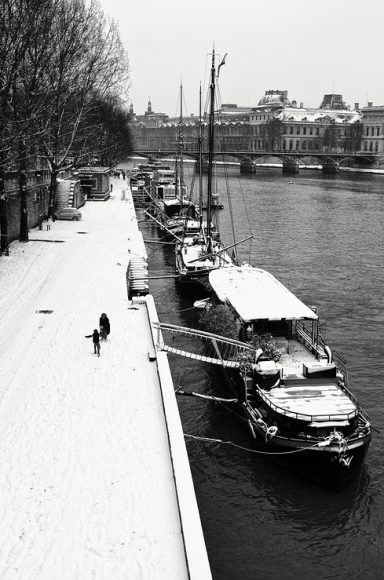 Black and white image of a snow-covered riverside with boats docked along the Seine River in Paris. A person with a dog walks along the snowy path, and trees line the bank to the left. In the background, buildings and a bridge crossing the river are visible, capturing Paris sous la neige.