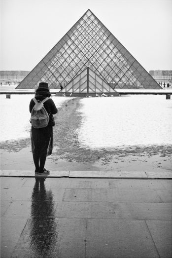A person wearing a hat and a backpack stands on a wet pathway, facing the glass pyramid of the Louvre Museum. Snow (or neige) covers the ground around the pathway. The scene is monochromatic, creating a stark, reflective atmosphere typical of a quiet samedi in winter.