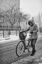 A person wearing a thick hooded parka, gloves, and boots is walking a bicycle along a snowy street one cold samedi. The scene includes snow-covered trees, a fence, and buildings in the background. The overall atmosphere is cold and wintry with gently falling neige.