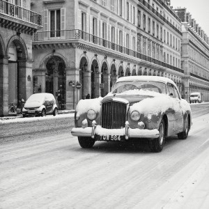 A vintage car covered in neige drives down a snow-covered street lined with elegant buildings with archways and balconies. Snow continues to fall, adding to the wintry atmosphere of this quiet dimanche morning. Other vehicles are parked along the street, also dusted with snow.